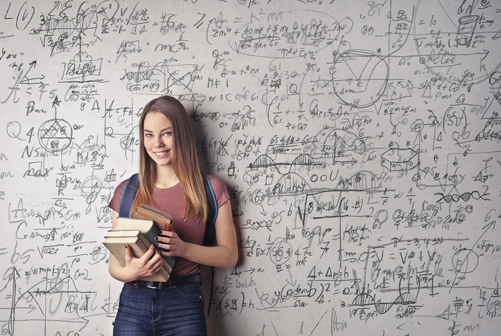 膨大な計算式などが書かれた白壁を背にハードカバーの本を数冊持って微笑む女性が立っている写真